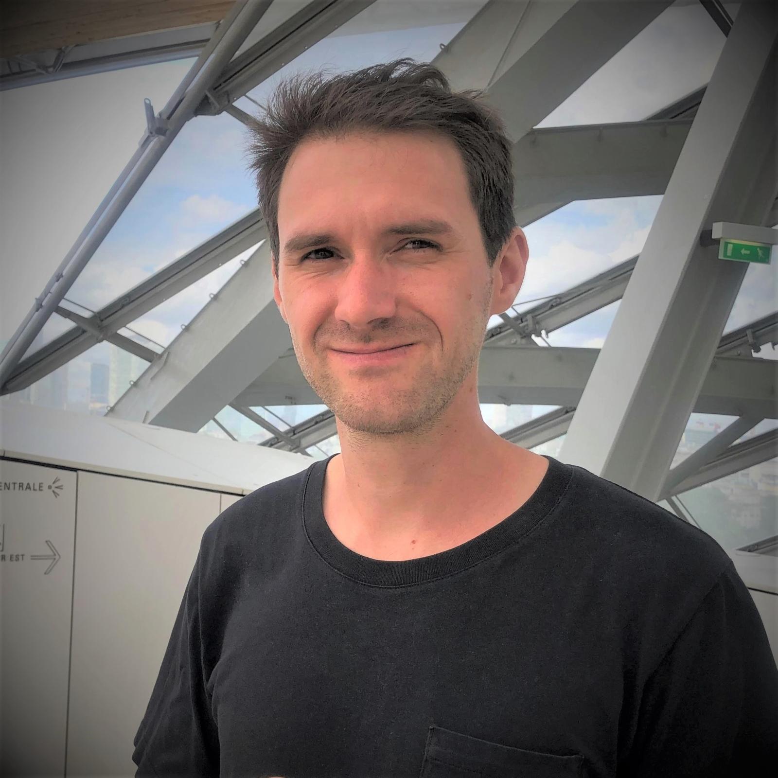 A picture of Vivien Butot, a man standing in front of a concrete building. He has dark hair and is wearing a dark grey t-shirt. 