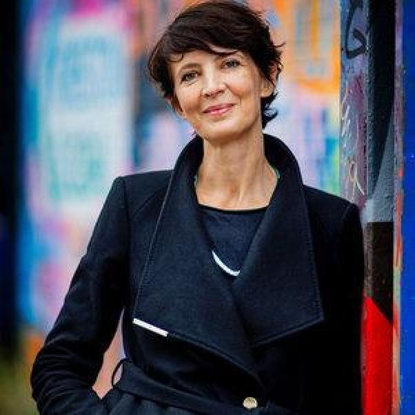 Bregje van Eekelen. A picture of a woman wearing a dark coloured coat. She has dark, short hair and is leaning against a wall covered in colour graffiti drawings.