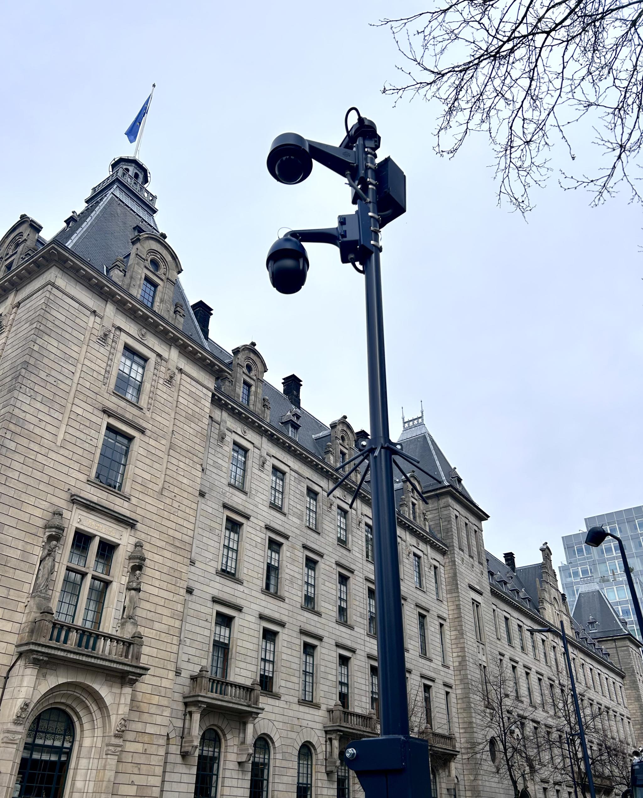 Een foto van een 'datapunt', een grote paal waar twee beveiligingscamera's aan gemonteerd zijn. Op de achtergrond het Rotterdamse stadhuis: een groot, statig gebouw met wit/zandkleurige bakstenen en een grijs dak.