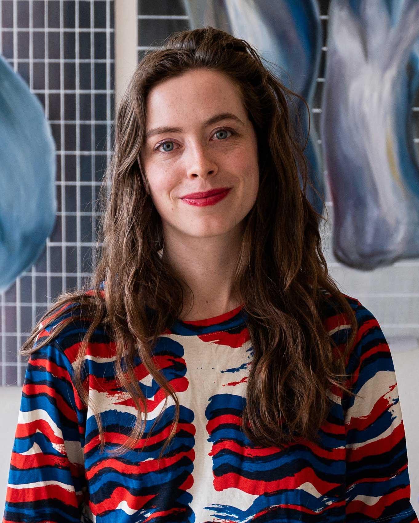 Gill Baldwin, a woman with long, dark hair standing in front of a large, blue-white and black painting. she is wearing a white blouse with red, blue and black stripes