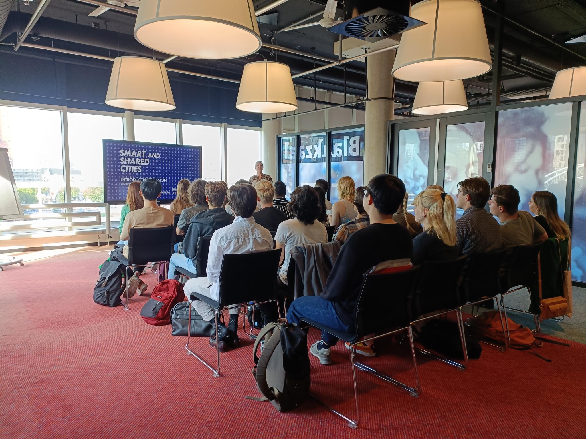 A group of students facing their teacher, in front of are large windows with a screen with a presentation in front of it