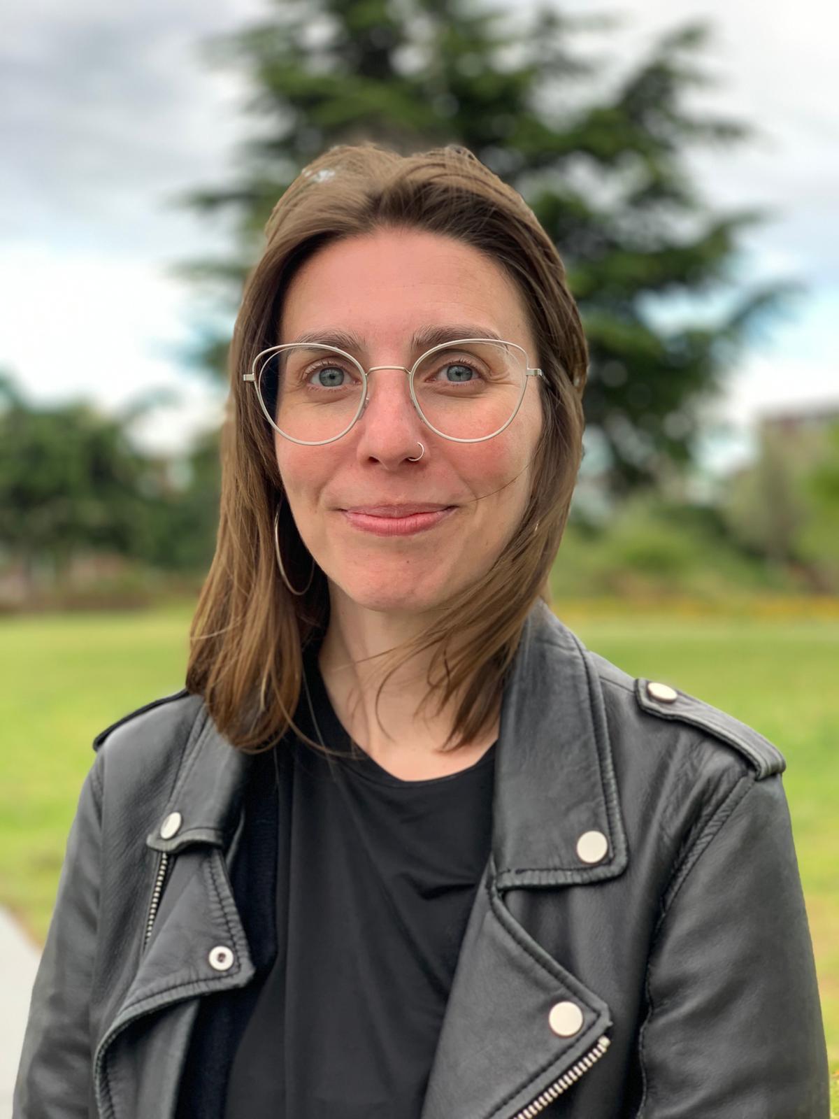 Isa Sanchez Cecilia. A picture of a smiling young women wearing a leather jacket. She has brown hair, glasses and a nose piercing and is standing in nature. 