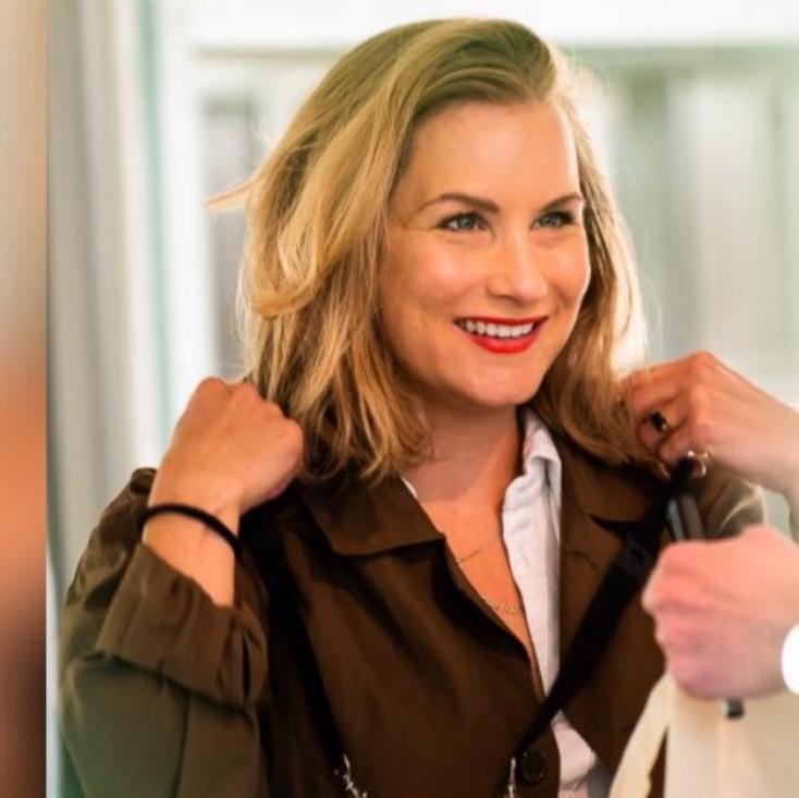 a photo of sabrinah huizenga. A woman, with long blonde hair who is smiling, while she is putting on a brown coat