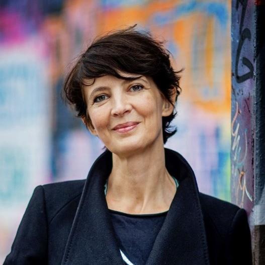 Bregje van eekelen. a woman with dark hair, smiling in the camera. She is standing in front of a multi-coloured background.