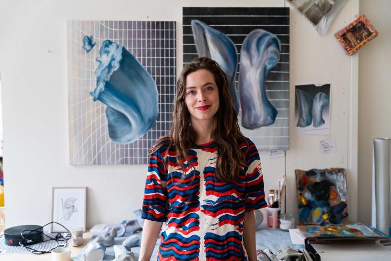 Gill Baldwin standing in her studio. She has dark hair and is smiling. She is wearing a red-white and blue blouse.