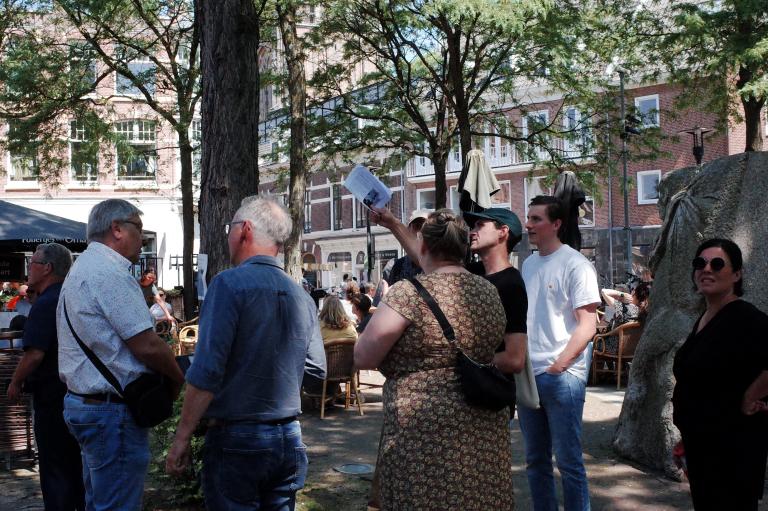 Group of people in discussion, one of them is pointing upwards. They are standing outside on a square. 
