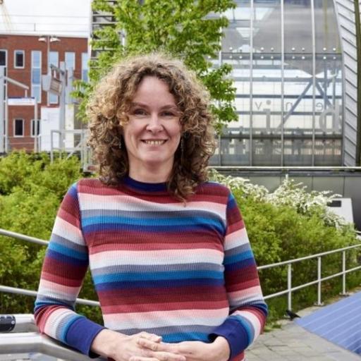 A picture of Marjolijn Das. She is standing on stairs, with greenery in the background. She has dark blond curly hair and is wearing a pink and blue striped sweater.