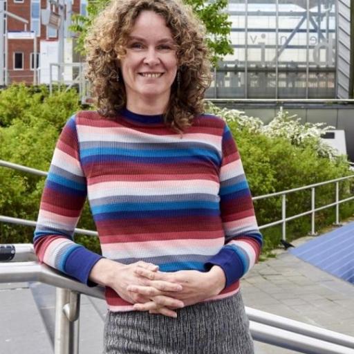 A picture of Marjolijn Das. She is standing on stairs, with greenery in the background. She has dark blond curly hair and is wearing a pink and blue striped sweater.