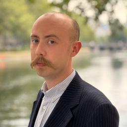 A picture of Leo Daly, a man standing in front of a natural background, dressed in a light blue shirt and black blazer